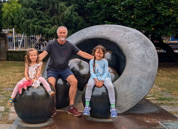 Jonny and his granddaughters sitting on large bronze apples with a giant bronze hat behind